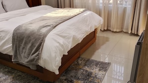 Ein ordentlich bezogenes Bett mit weißer Daunendecke und grauer Tagesdecke, ein Holz-Kopfteil und ein gemustertes Teppich auf Fliesen.