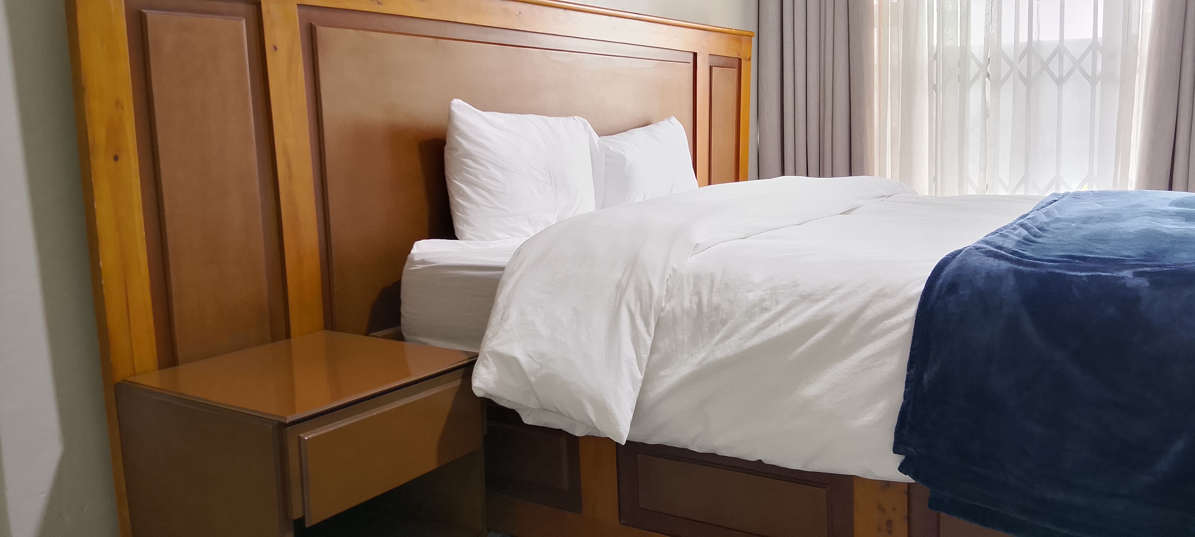 Habitación de hotel con cabecera de madera, cama individual con ropa blanca, mesita de noche y ventana con cortinas.