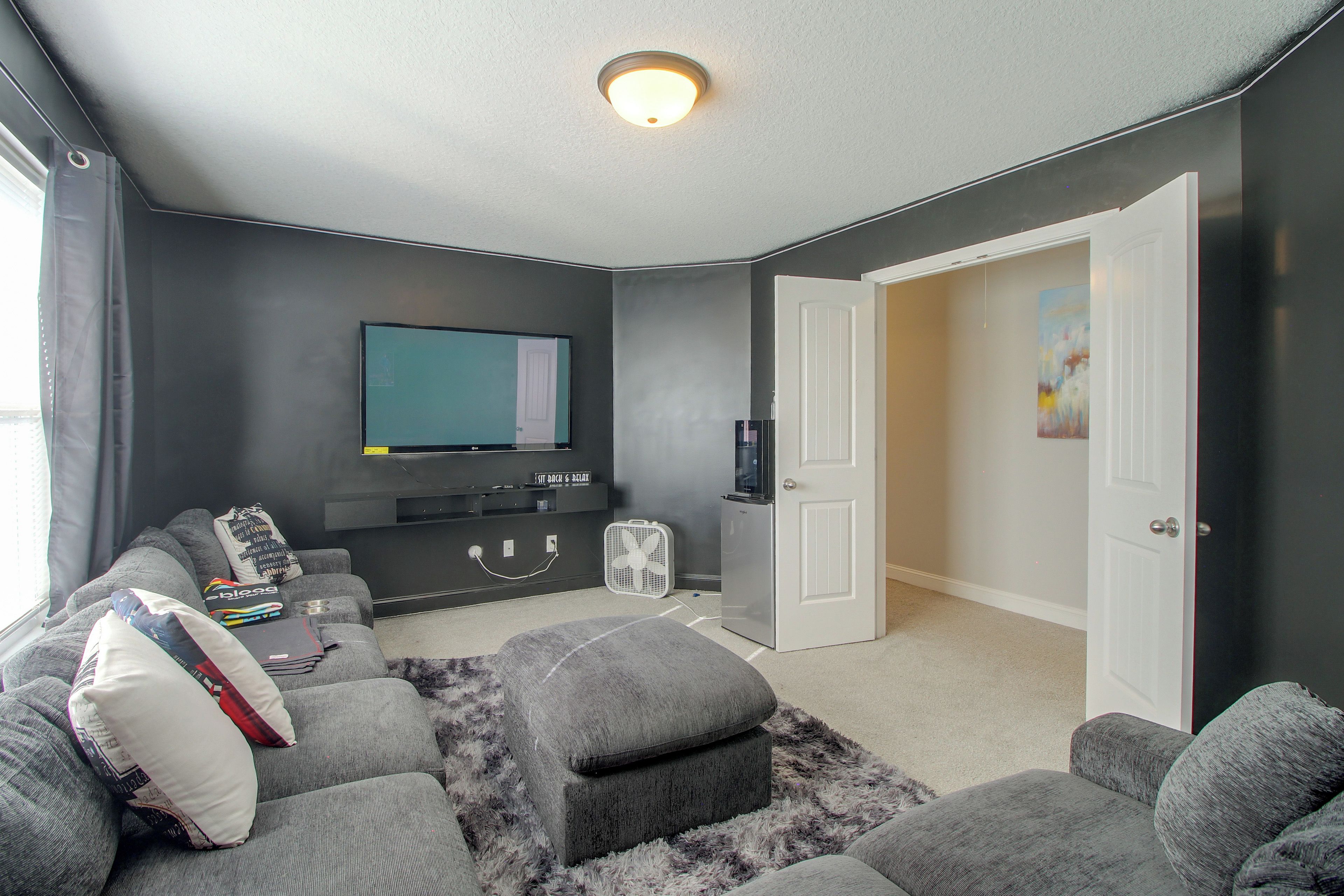 A living room with a flat-screen TV, grey sofas, a small white fridge, and a fluffy rug.