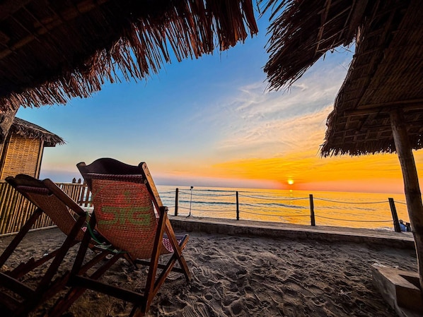 A beach view with a sunset, a wooden chair, and a thatched roof structure.