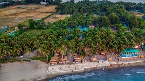 Vista aérea de una playa con palmeras, una orilla arenosa y un edificio con techo azul.