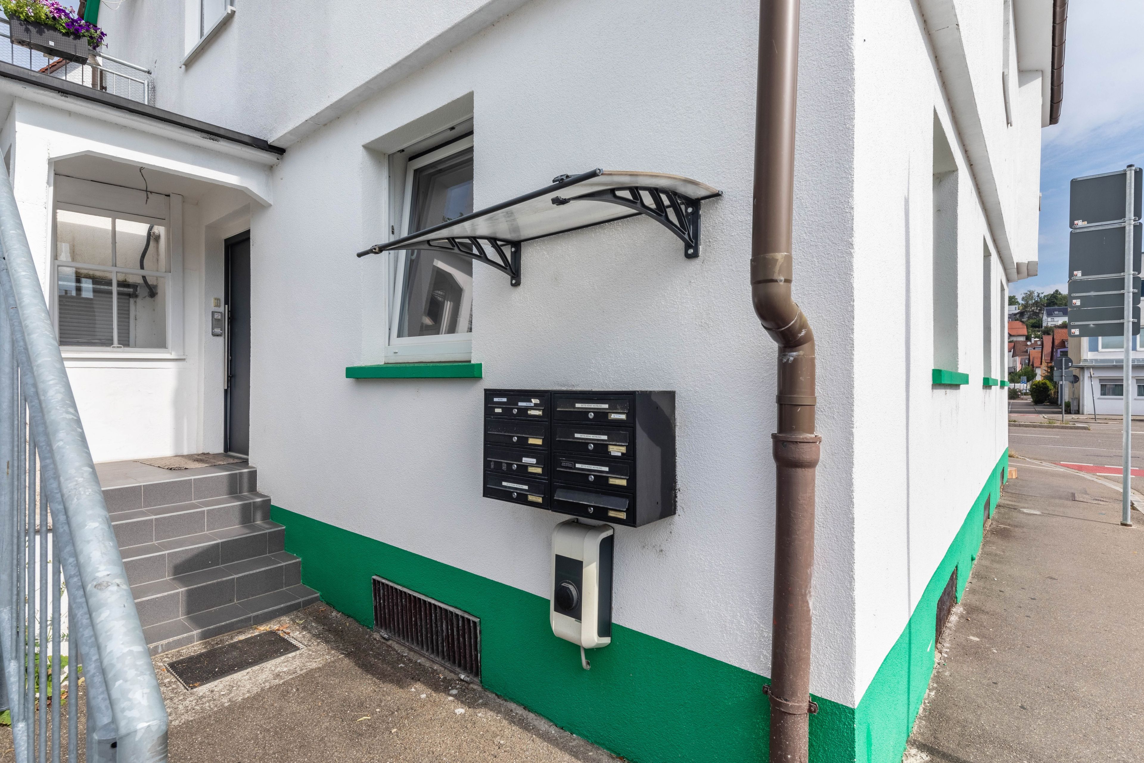 Ein Gebäude mit weißer und grüner Fassade, einer Metalltreppe, einem Briefkasten und einer Überwachungskamera.