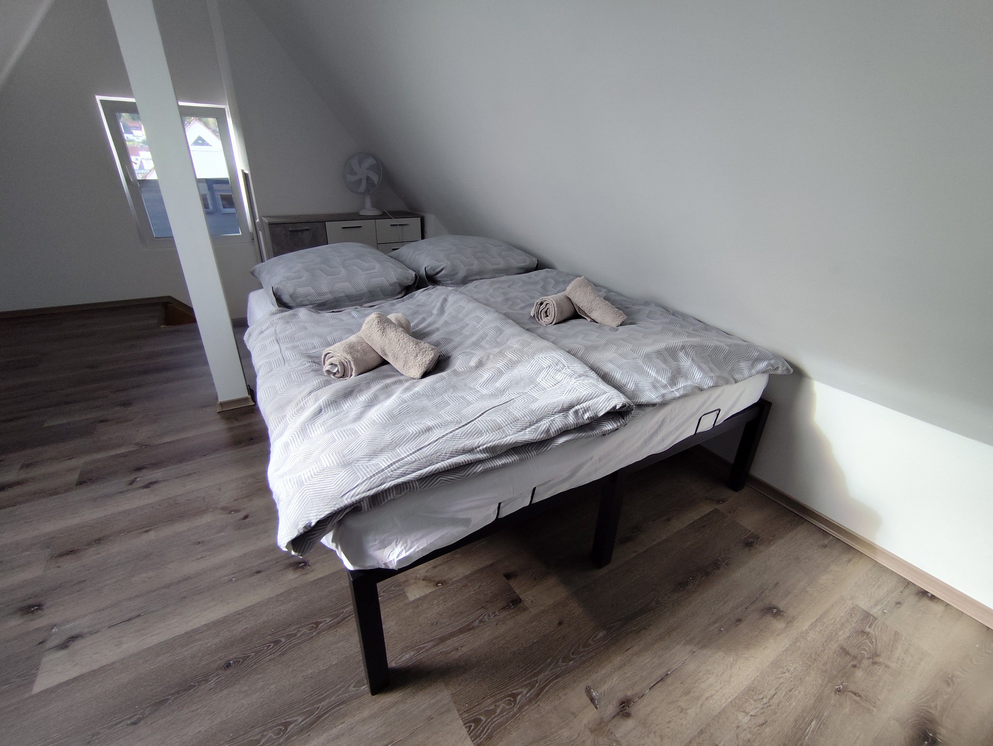 Un lit simple avec une couvre-lit à motifs et deux serviettes enroulées, une table de chevet avec un ventilateur et un plancher en bois.