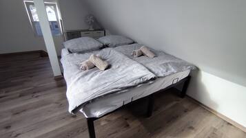 Un lit simple avec une couvre-lit à motifs et deux serviettes enroulées, une table de chevet avec un ventilateur et un plancher en bois.