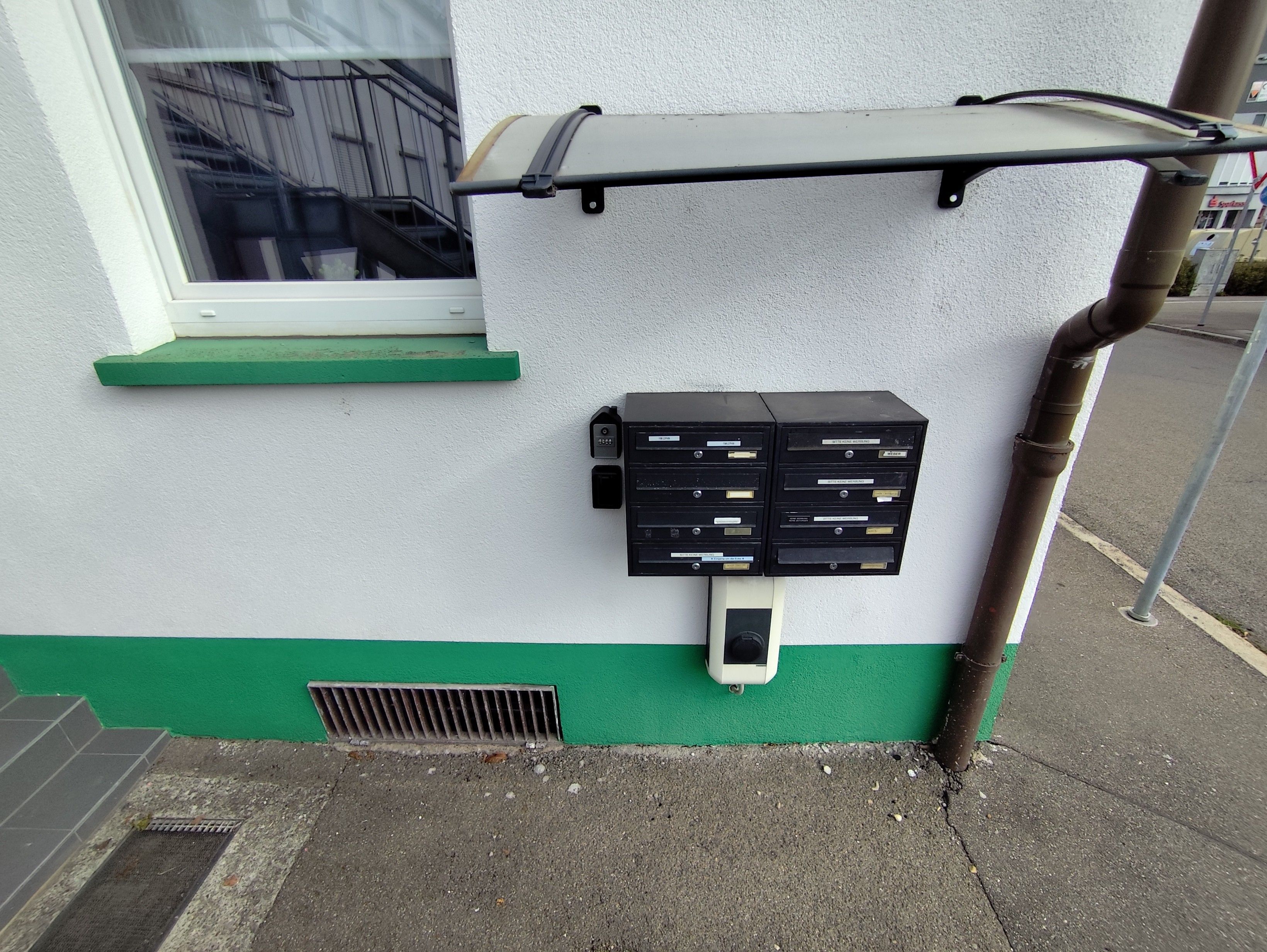 A utility box with a green base and a white wall, next to a window with a metal guard.
