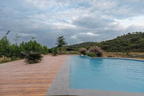 A swimming pool with a tiled deck, surrounded by greenery and hills in the background.