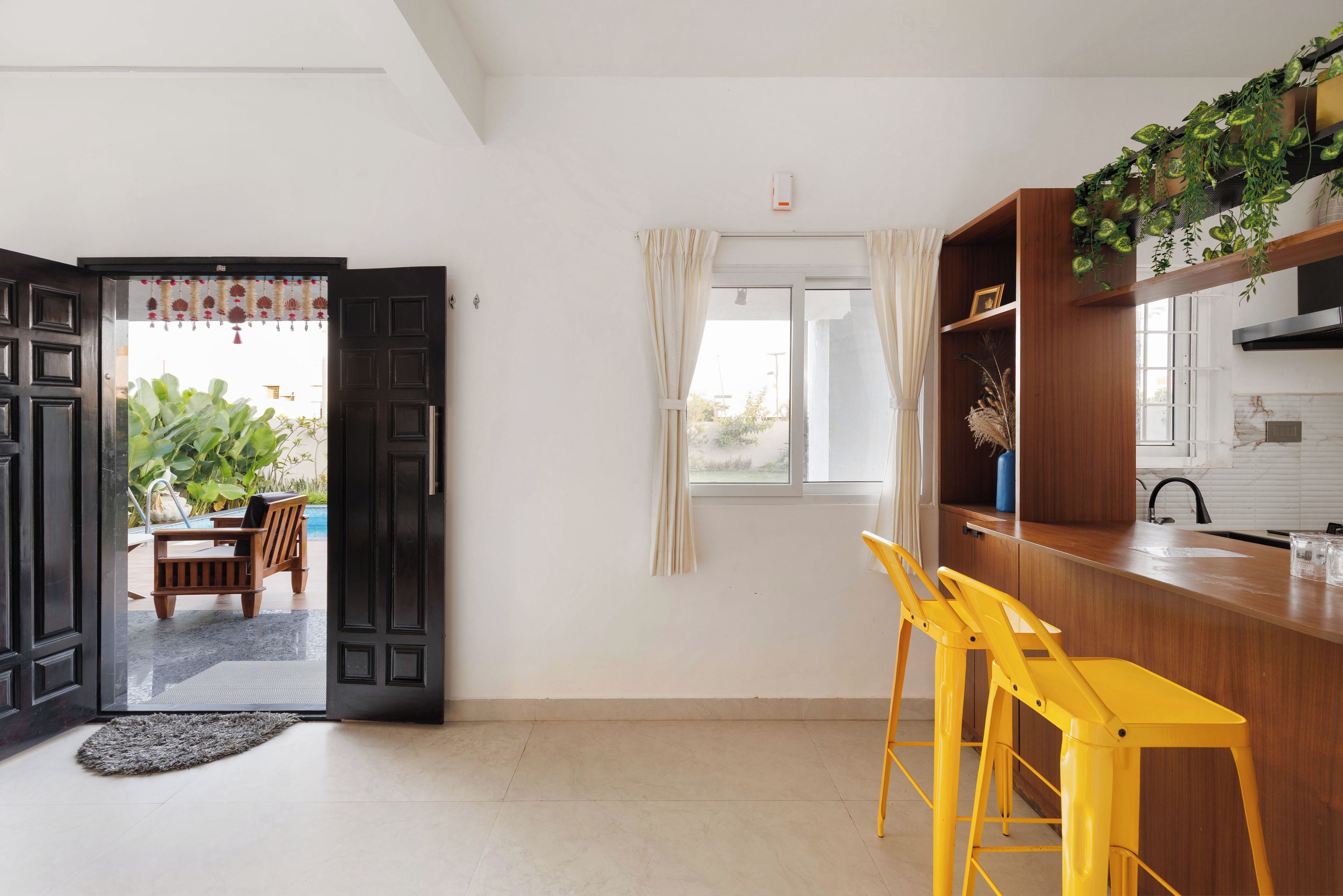 A modern kitchen with wooden cabinets, a bar area with yellow stools, and a view through the open door to an outdoor seating area.