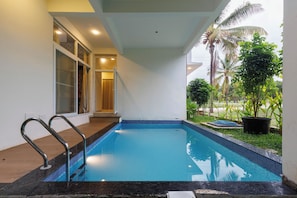 A swimming pool with a tiled edge, a metal ladder, and a glass door leading to a covered area. - Sierramystique by Stayjadepool3bhk35min Airport (Doddaballapur)