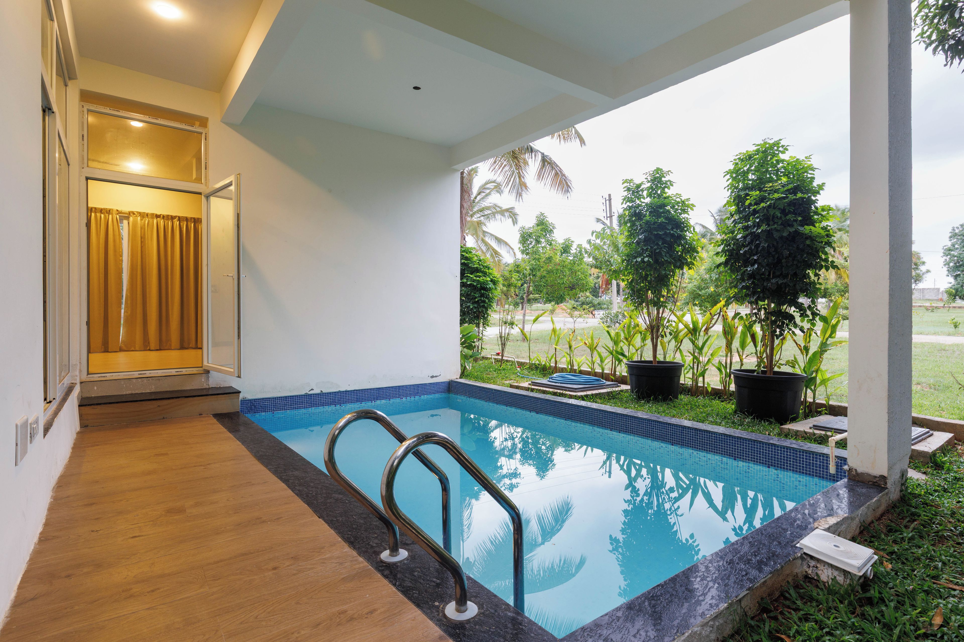 A swimming pool with a wooden deck and metal handrails, surrounded by potted plants and a view of a grassy area.