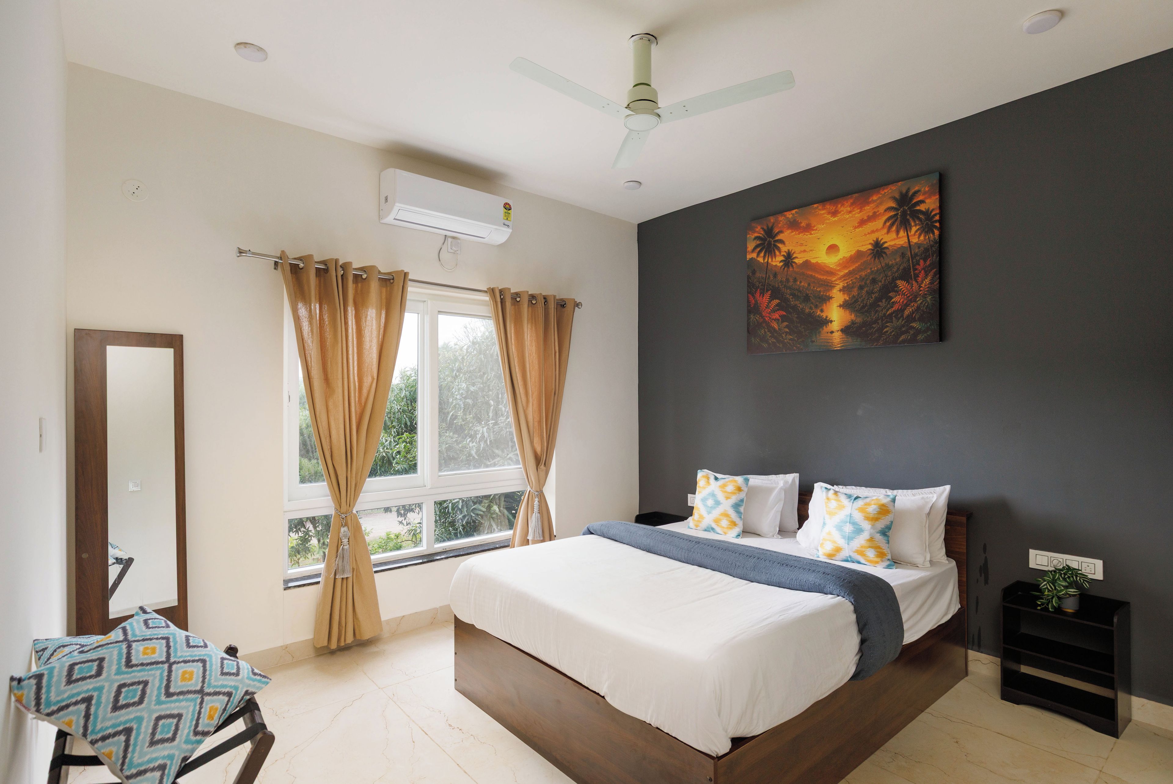 A bedroom with a bed, curtains, a ceiling fan, a painting, and a window with a view of greenery.