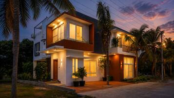 A two-story modern house with a balcony, surrounded by palm trees and a well-maintained garden.