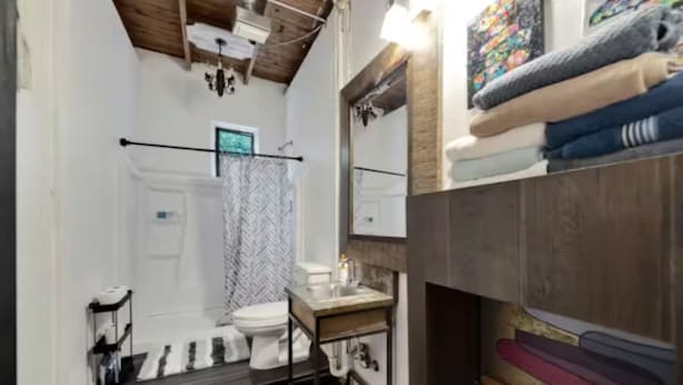 A bathroom with a wooden ceiling, a white toilet, a sink with a mirror, and a towel rack with folded towels. - Art Studio in DOWNTOWN Channelside (Tampa)