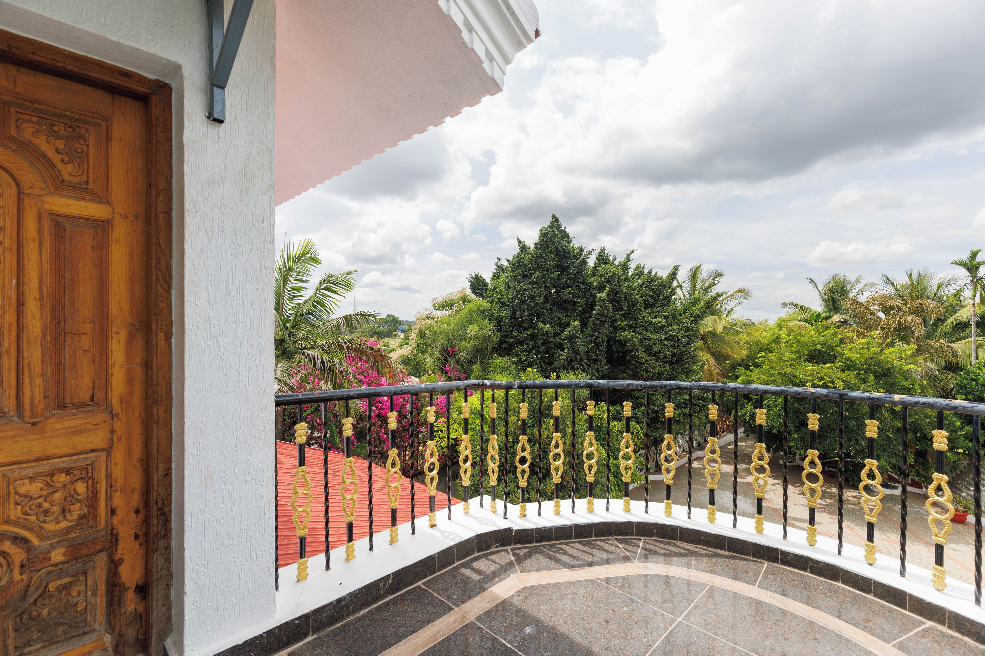 A balcony with a curved railing, a wooden door, and a view of a garden with various plants and a red-roofed structure.