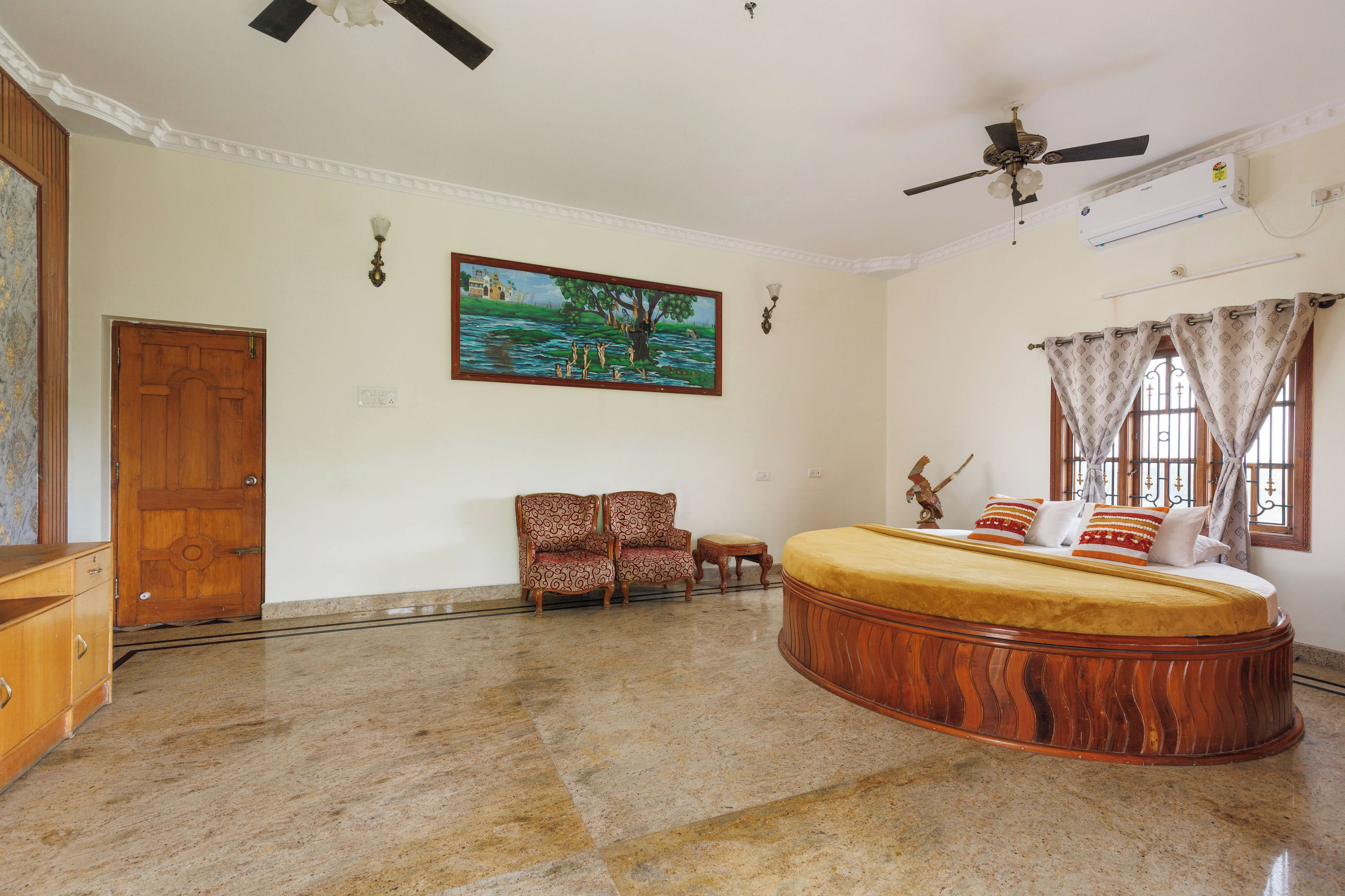 A spacious bedroom with a large bed, a ceiling fan, an air conditioning unit, a painting on the wall, and a window with curtains.