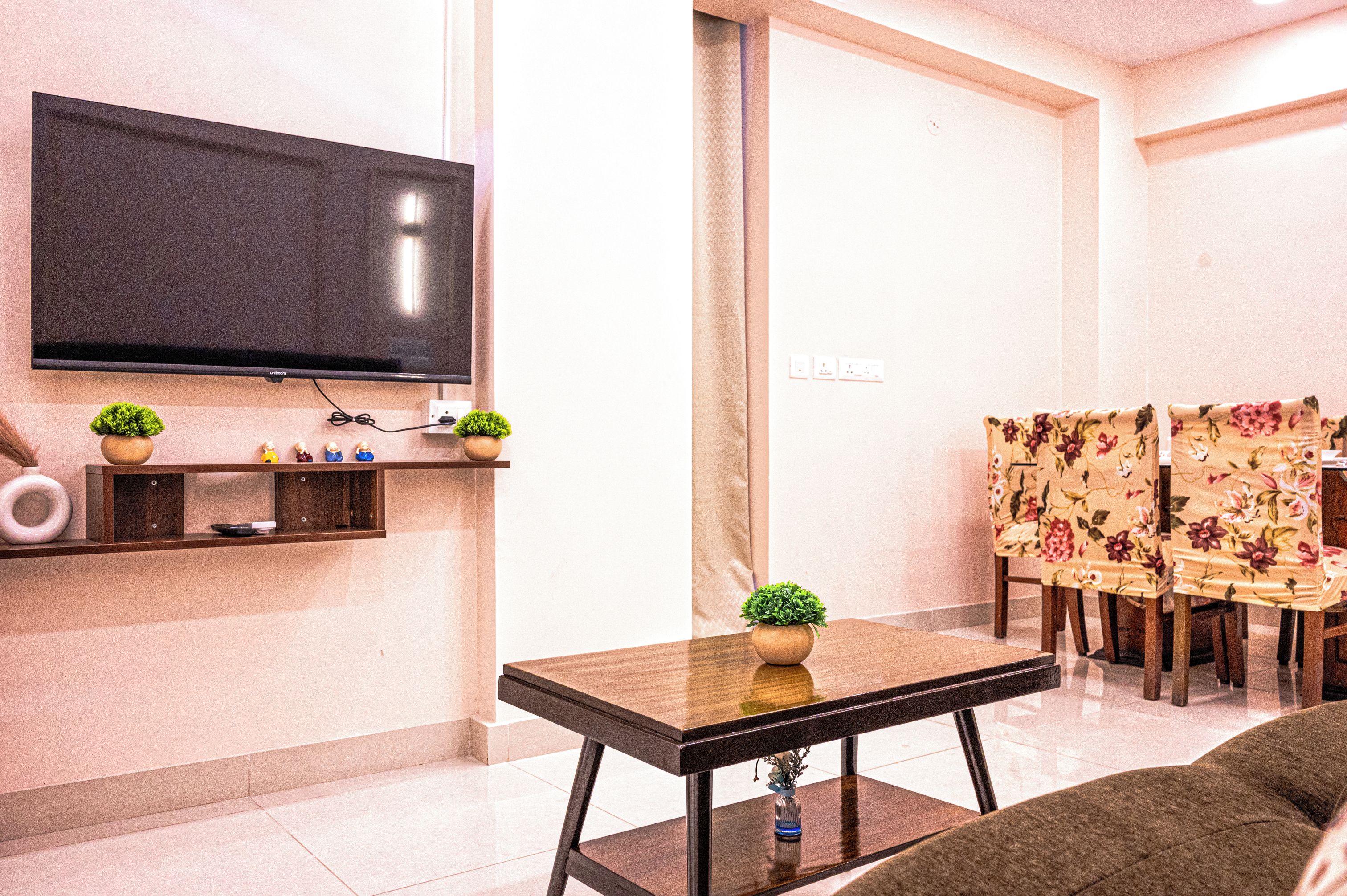 A modern living room with a flat-screen TV mounted on the wall, a wooden coffee table with a potted plant, a sofa, and a floral-patterned dining table with chairs.