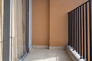 A narrow outdoor corridor with a tiled floor, a glass door, and a black metal railing.