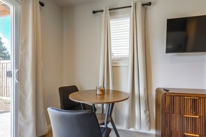 Ein Zimmer mit einem runden Holztisch, zwei Stühlen, einem an der Wand befestigten Fernseher und einem Fenster mit Jalousien.