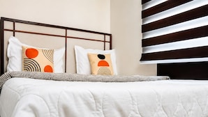 Un lit bien fait, avec une couette blanche et des oreillers à motifs, une tête de lit à motifs géométriques et une fenêtre avec des stores horizontaux.