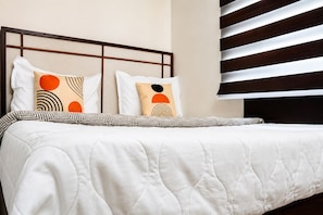 A neatly made bed with a white quilt and patterned pillows, a headboard with a grid design, and a window with horizontal blinds. - The Icon by Redolive 2bhkkoyambeduit Parkdlf (Chennai)