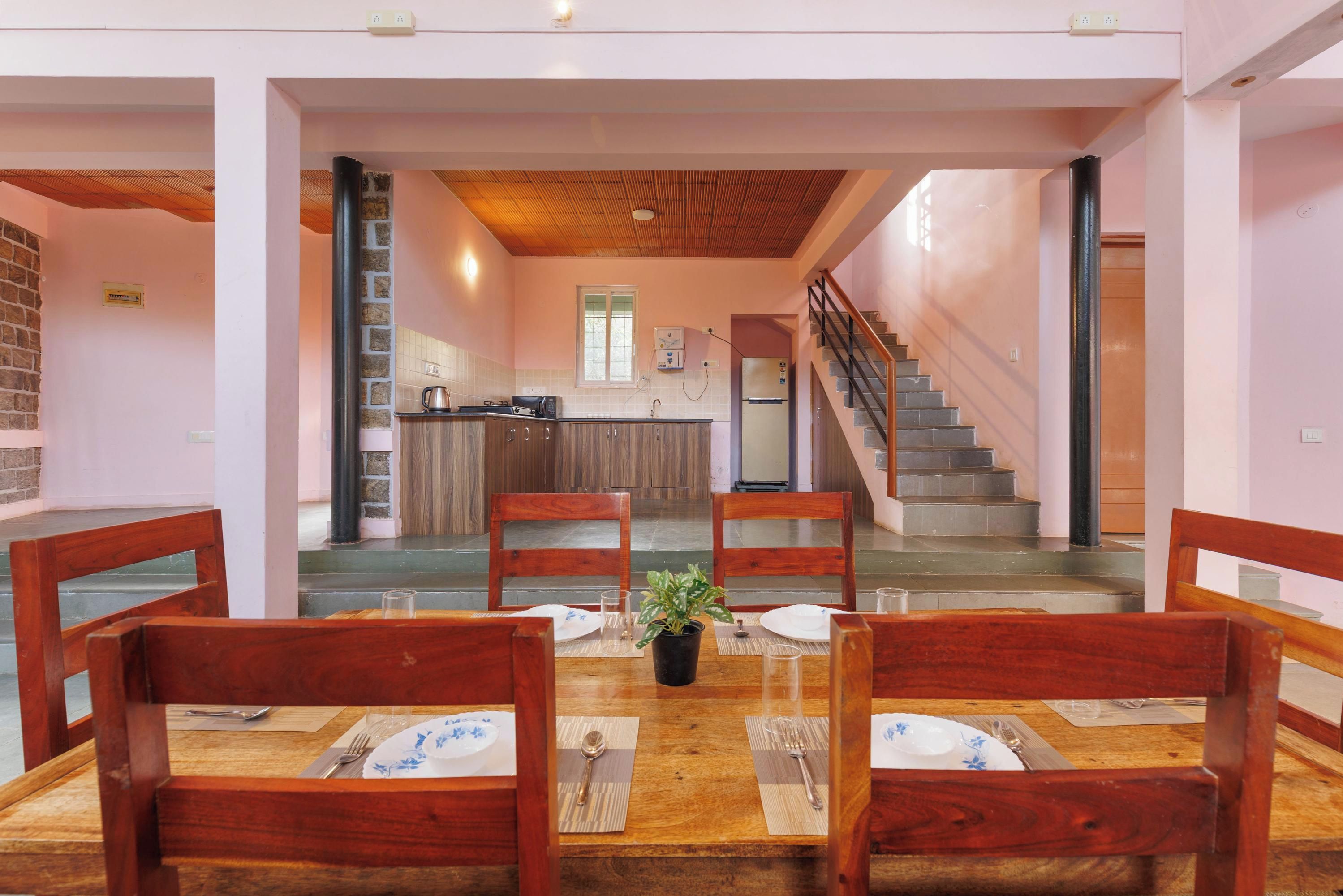 A dining area with wooden chairs and a table set for four, a potted plant, and a kitchen in the background.