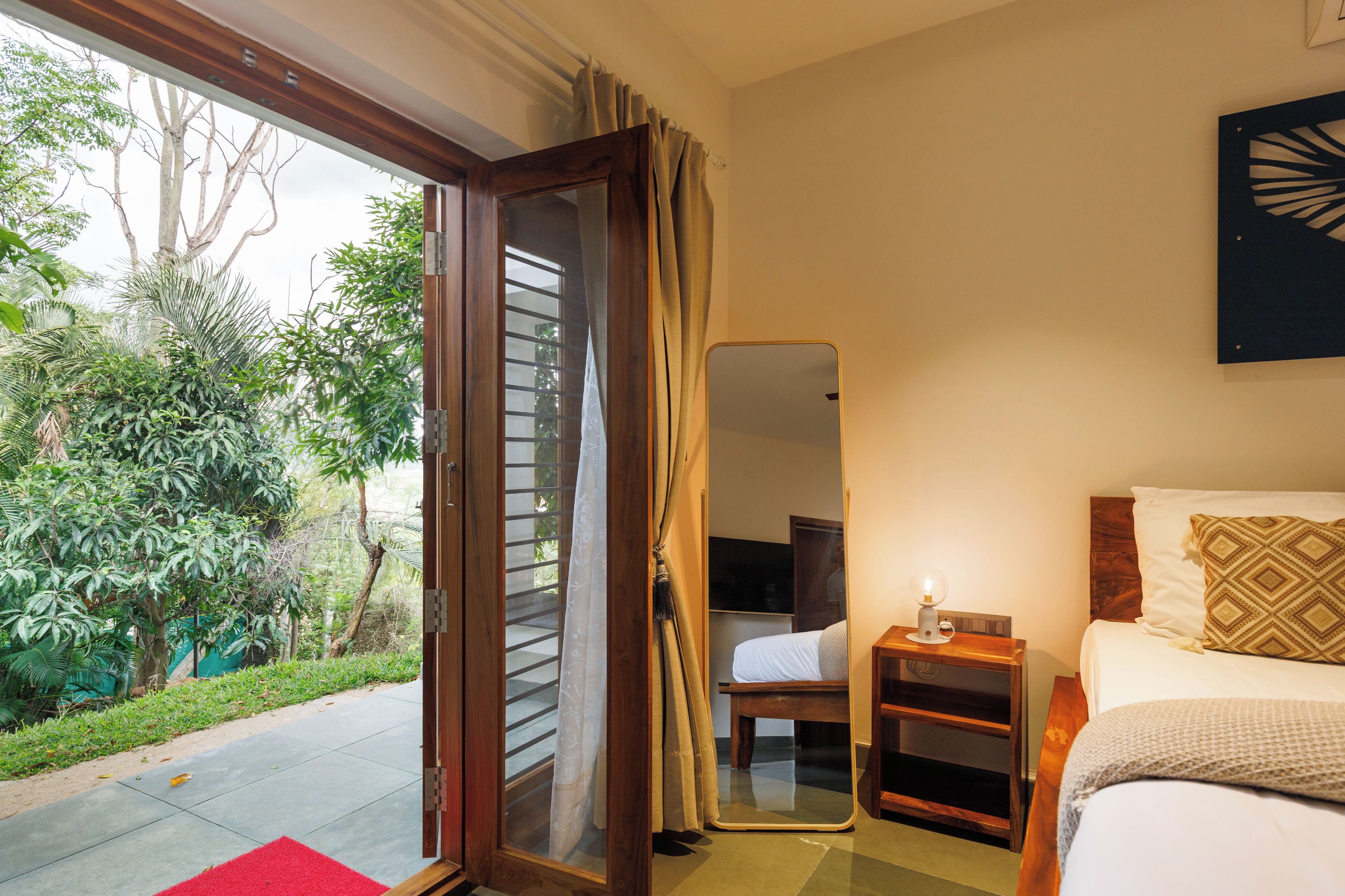 Un dormitorio con cama, mesita de noche, espejo y vista a un jardín por una puerta abierta.