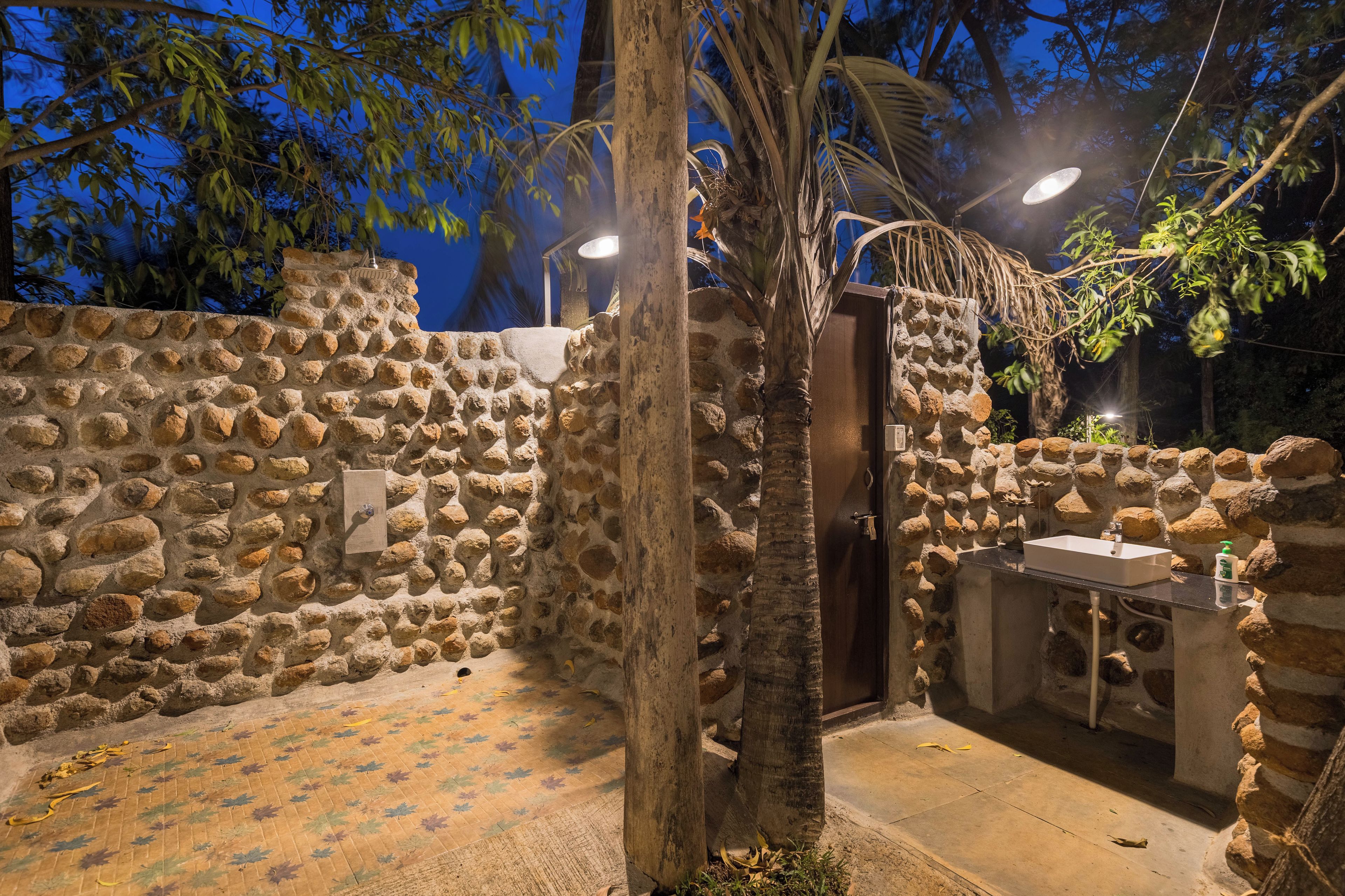 Un área de ducha al aire libre con paredes de piedra, una puerta de madera, piso de baldosas y un pequeño lavamanos con espejo.