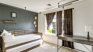 Une chambre à coucher avec un lit, un bureau, une fenêtre avec des rideaux, un miroir fixé au mur et un cadre photo.