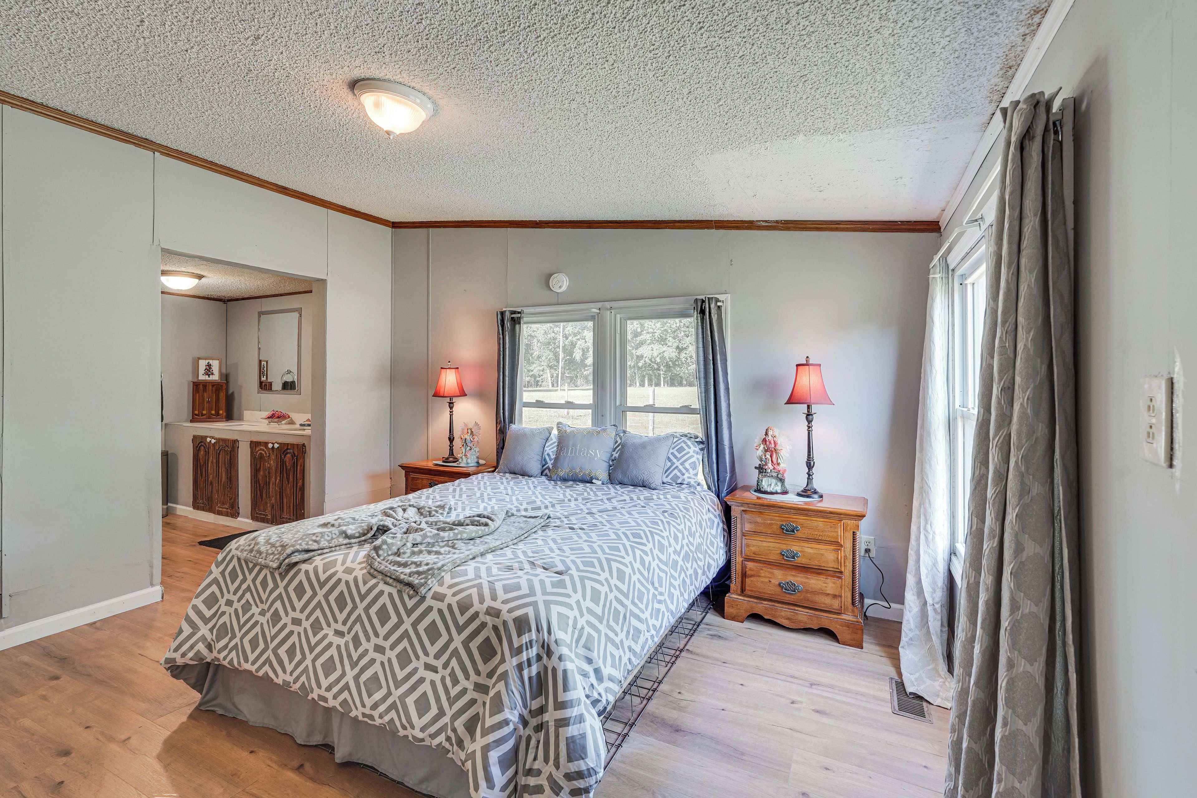 A bedroom with a bed, two bedside tables with lamps, a window with curtains, a mirror, and a wooden dresser.