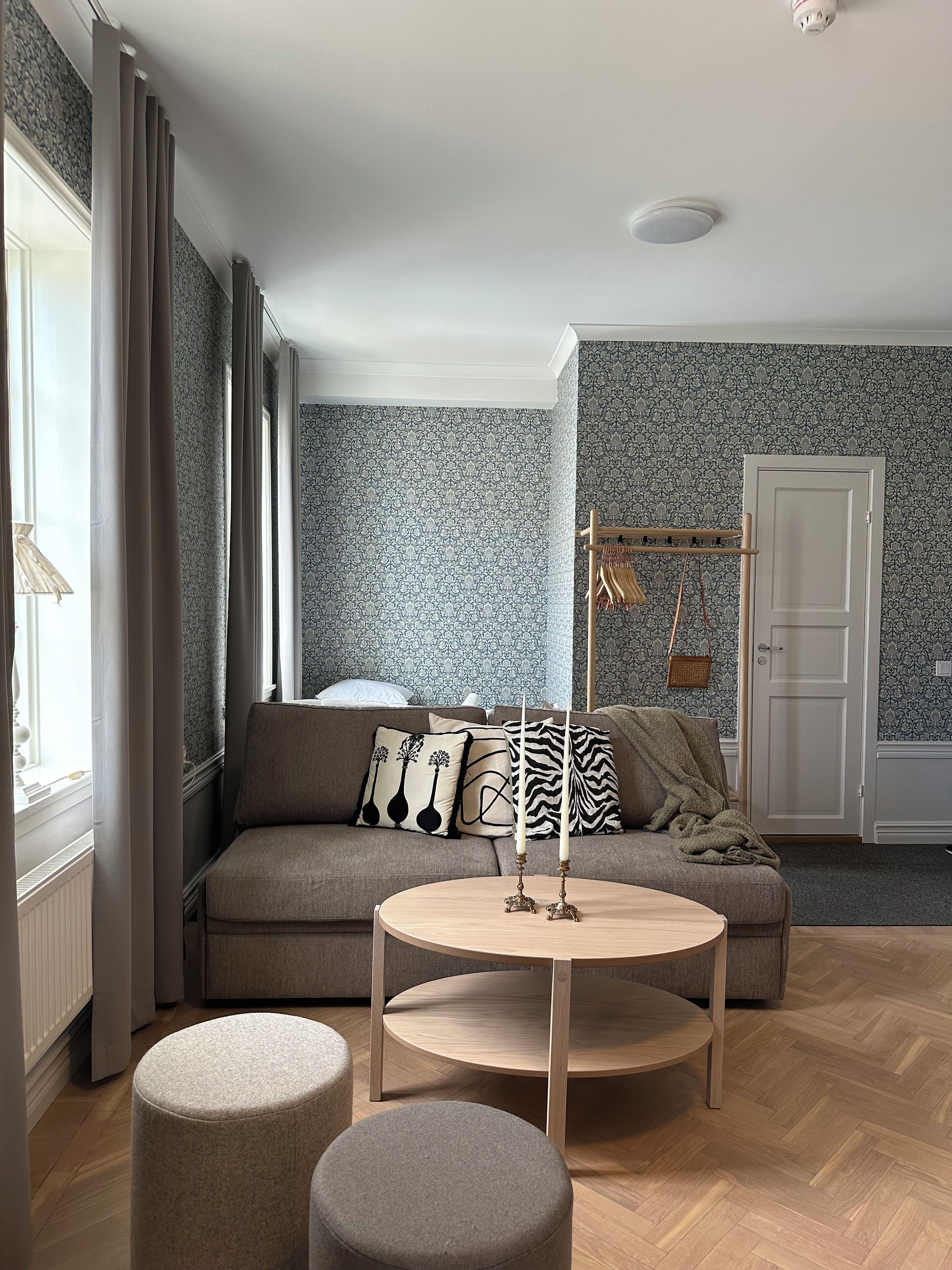 A living room with a sofa, two ottomans, a round coffee table, patterned wallpaper, a door, and a coat rack.