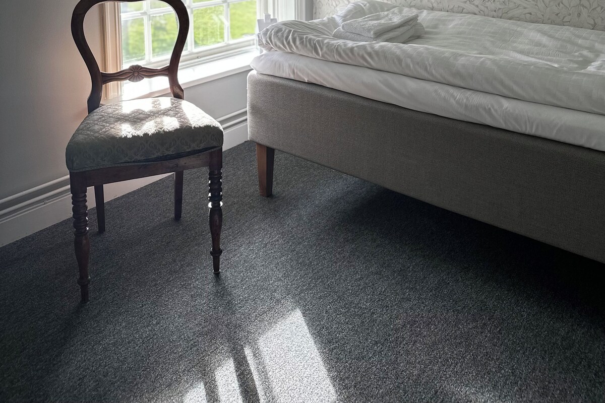 Quarto com uma cama de solteiro, uma cadeira, uma janela e luminária fixada na parede.