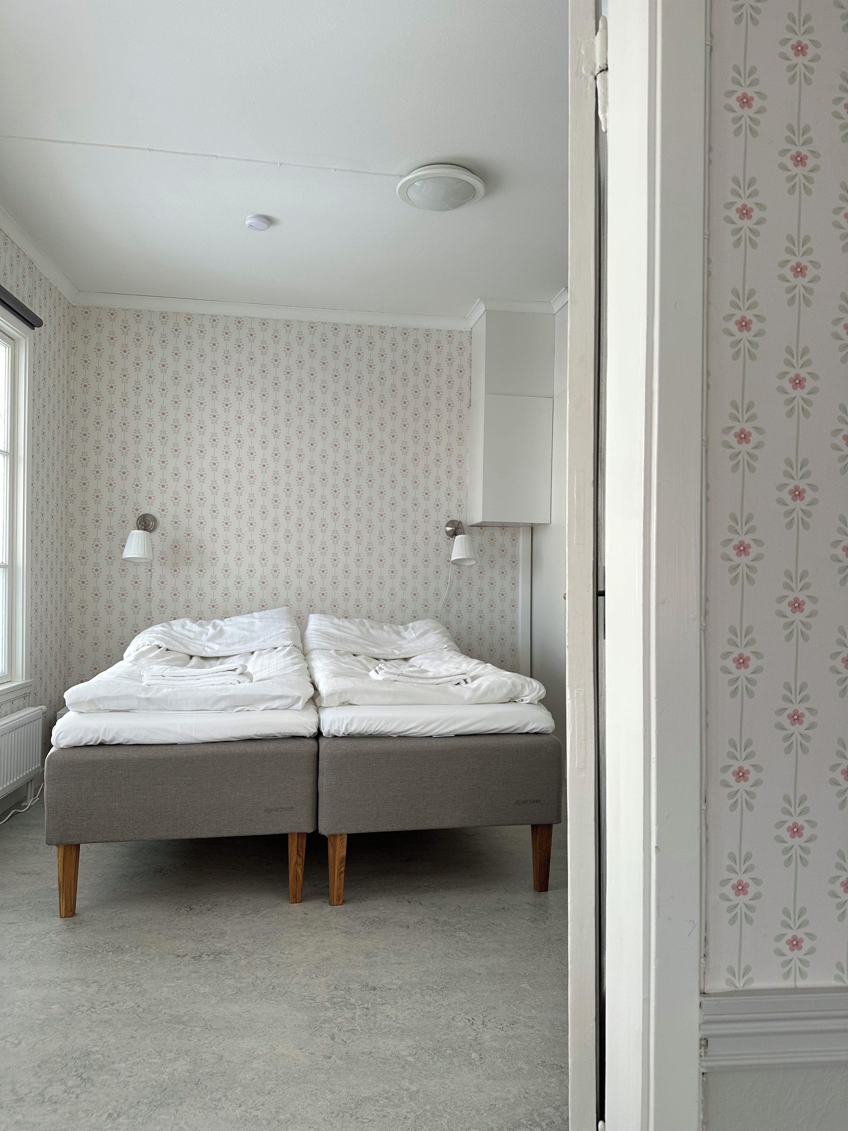 Un dormitorio con una cama, dos lámparas de noche, un radiador y papel tapiz con motivos florales.