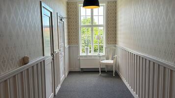 En gang med tapet med mønster, en radiator, et vindue med udsigt over grønne områder og en pendellampe.