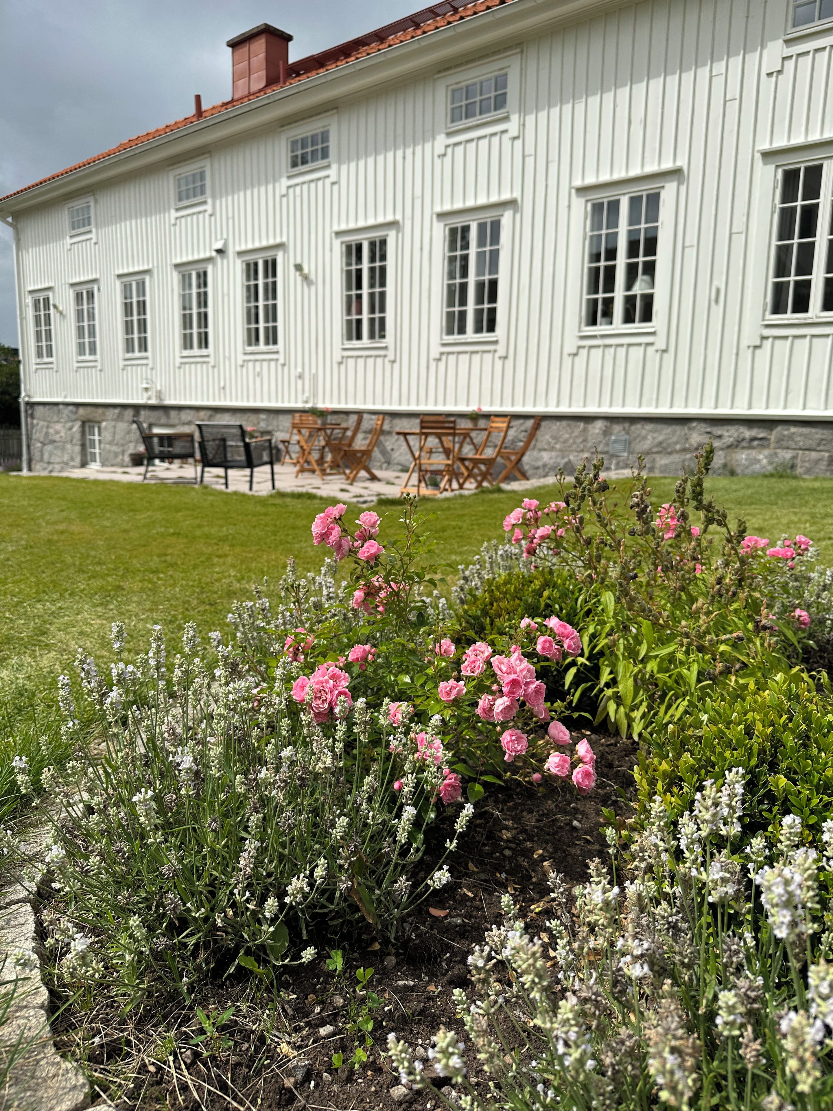 Um edifício branco com várias janelas e um telhado vermelho, rodeado por um jardim bem cuidado com flores cor-de-rosa e brancas.