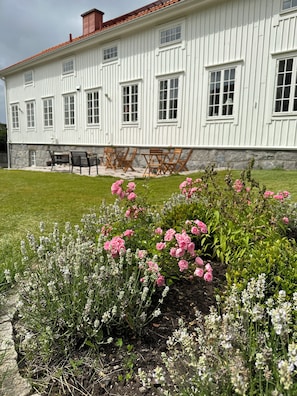 Un edificio blanco con múltiples ventanas y un techo rojo, rodeado de un jardín bien cuidado con flores rosas y blancas.
