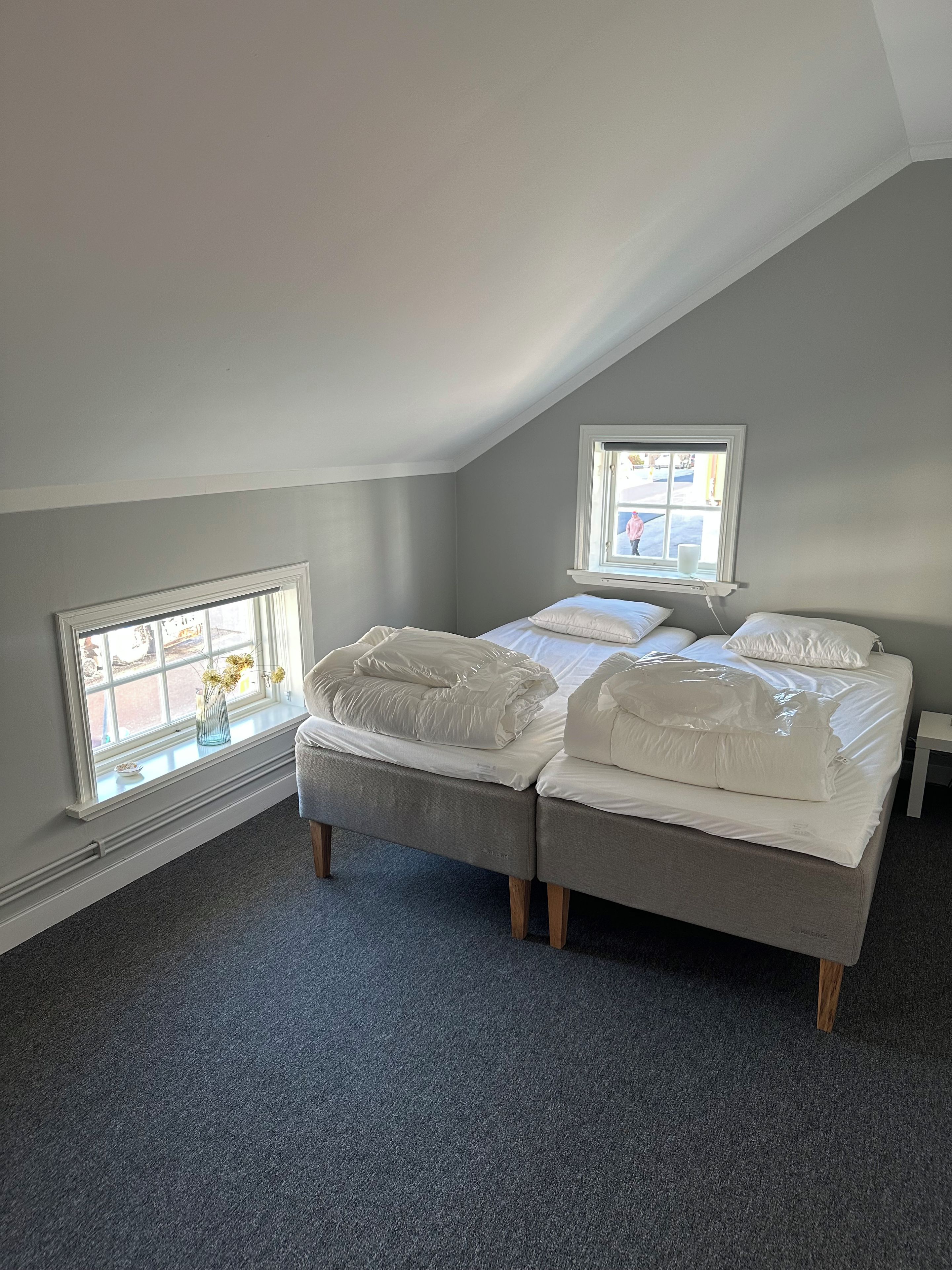Un dormitorio con techo inclinado, dos camas con ropa de cama blanca, una ventana con vista a la calle y una mesita.