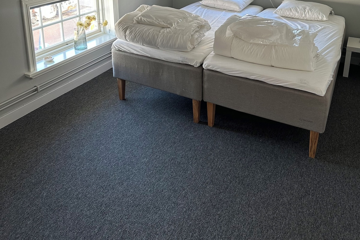 Un dormitorio con techo inclinado, dos camas con ropa de cama blanca, una ventana con vista a la calle y una mesita.