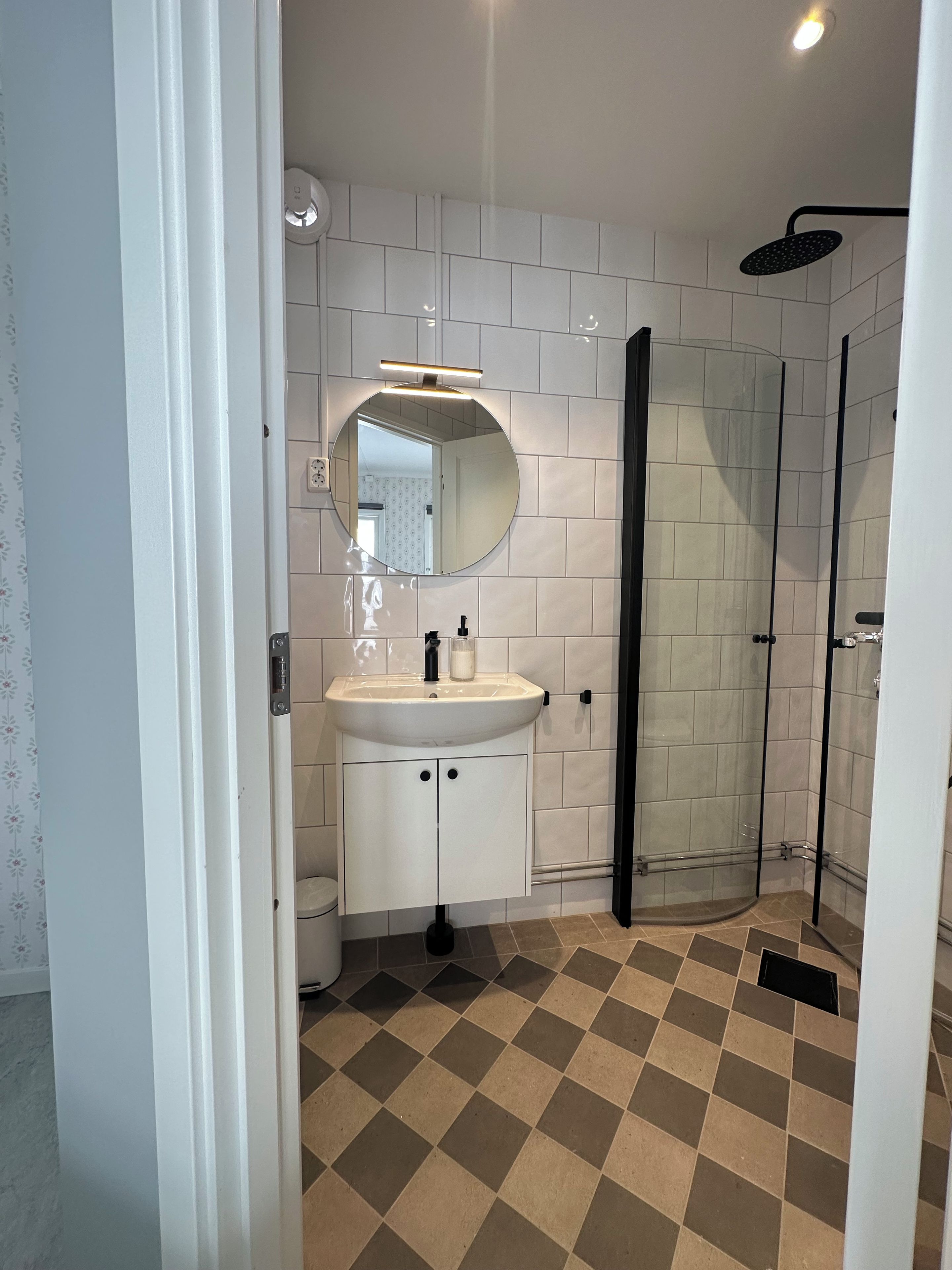 A bathroom with a white tiled wall, a round mirror, a sink with a black faucet, and a shower area with a glass door.