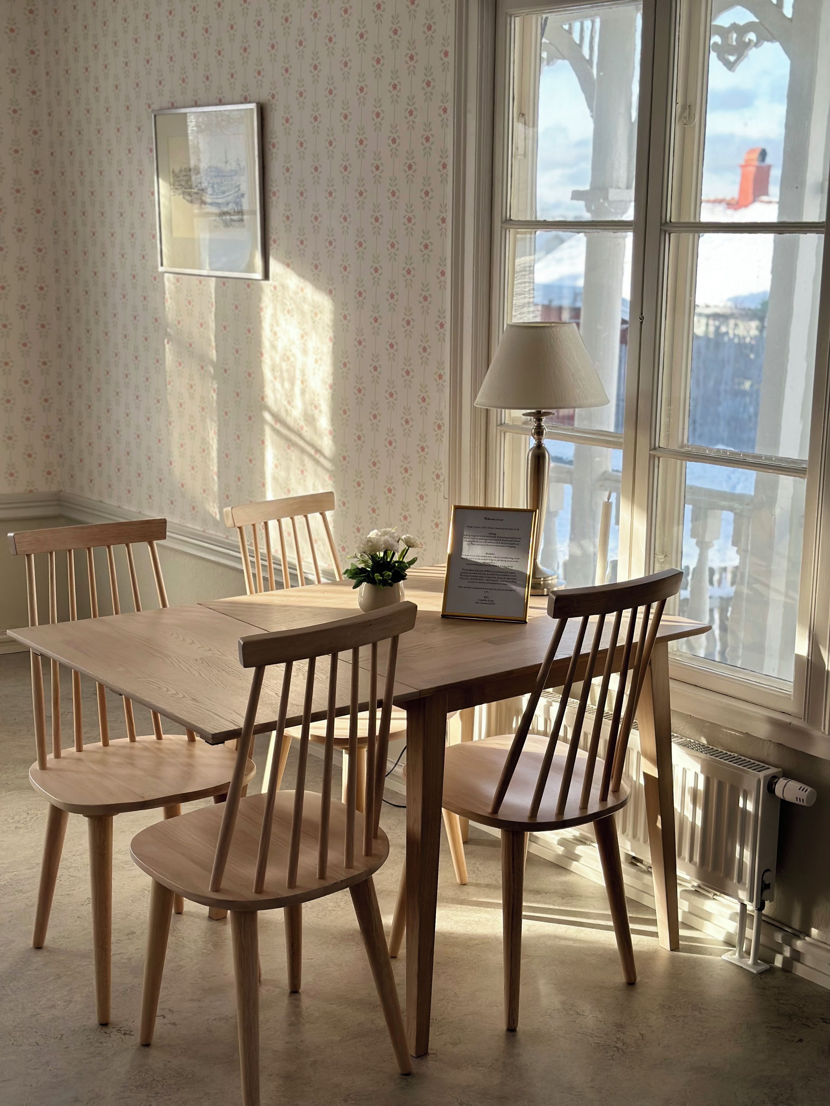 Un coin repas avec une table et des chaises en bois, un certificat encadré, une plante en pot et une fenêtre donnant sur des immeubles.