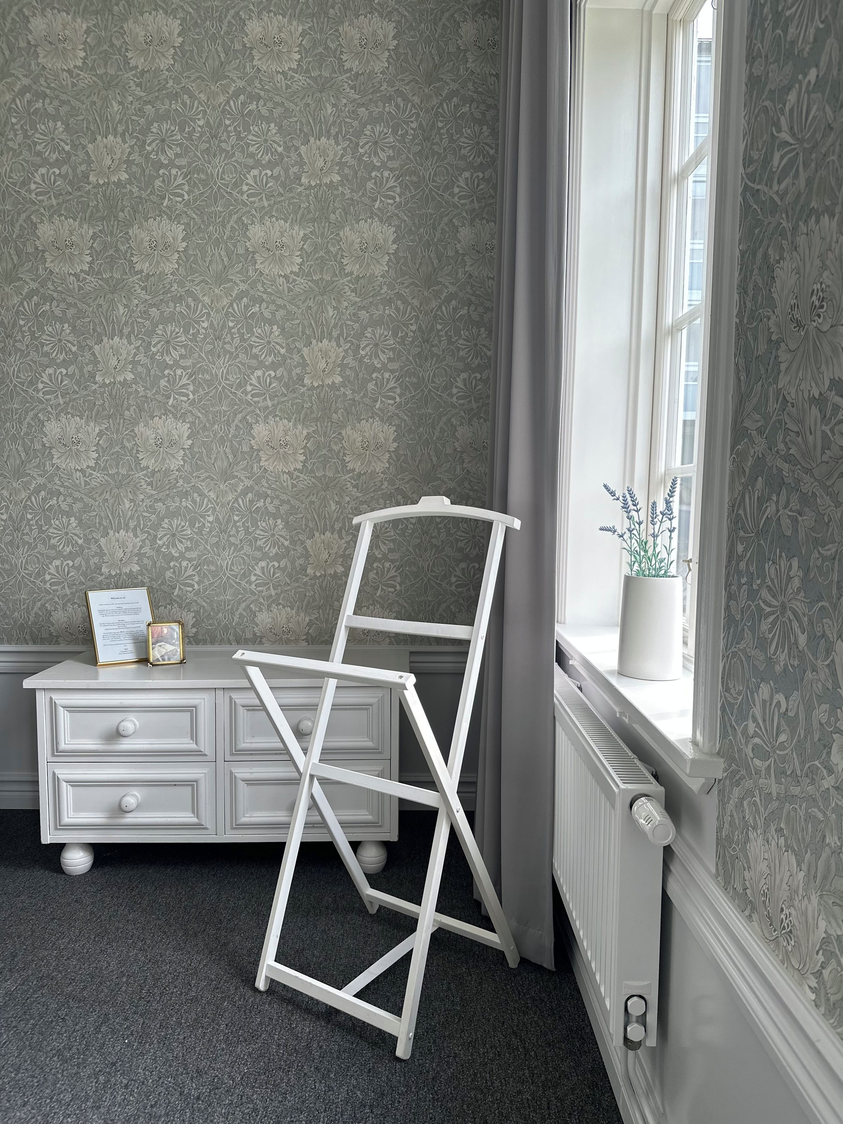 A room with a radiator, a white dresser with drawers, a framed certificate, and a white ladder leaning against the wall.