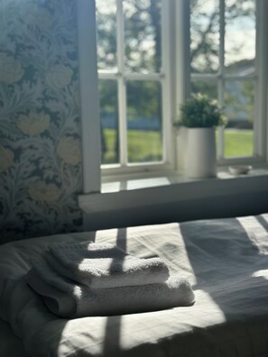 Una cama con sábana blanca y una toalla doblada, una ventana con vistas a la vegetación y una planta en maceta sobre el alféizar.