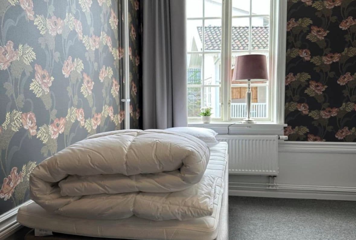 A neatly made bed with a white comforter and pillows, a window with a view of a house, and floral wallpaper.