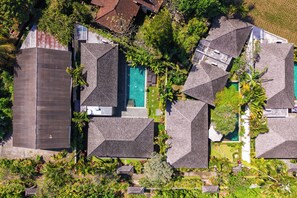 Vista aérea de un barrio residencial con varias casas, cada una con un diseño de techo diferente, rodeado de vegetación exuberante y una piscina.