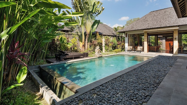 Piscina com borda de pedra, rodeada por plantas tropicais e um edifício com telhado em azulejos.
