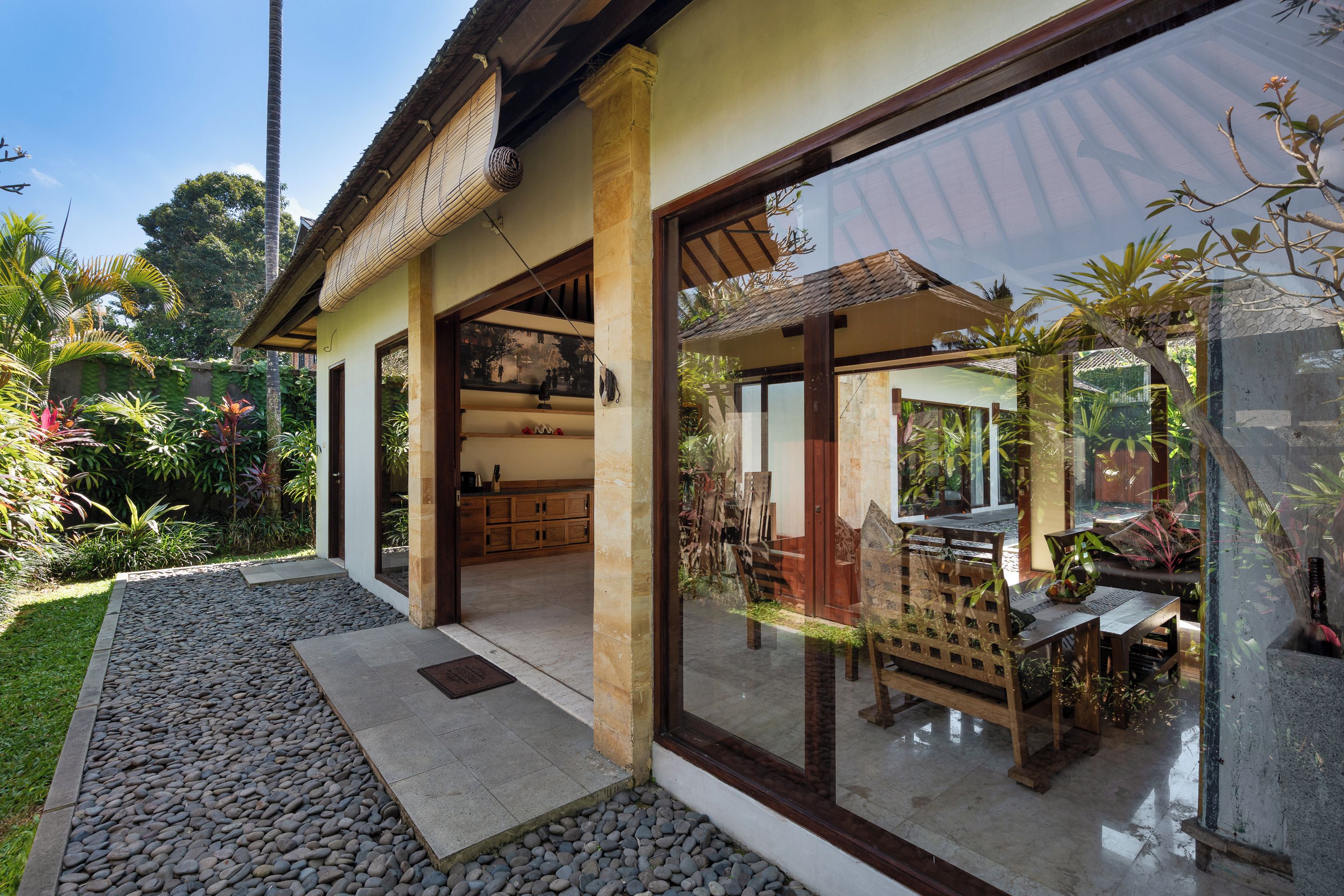 Una casa moderna con un amplio ventanal de vidrio que da a un comedor al aire libre, rodeada de plantas tropicales.