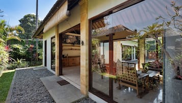 Una casa moderna con un amplio ventanal de vidrio que da a un comedor al aire libre, rodeada de plantas tropicales.