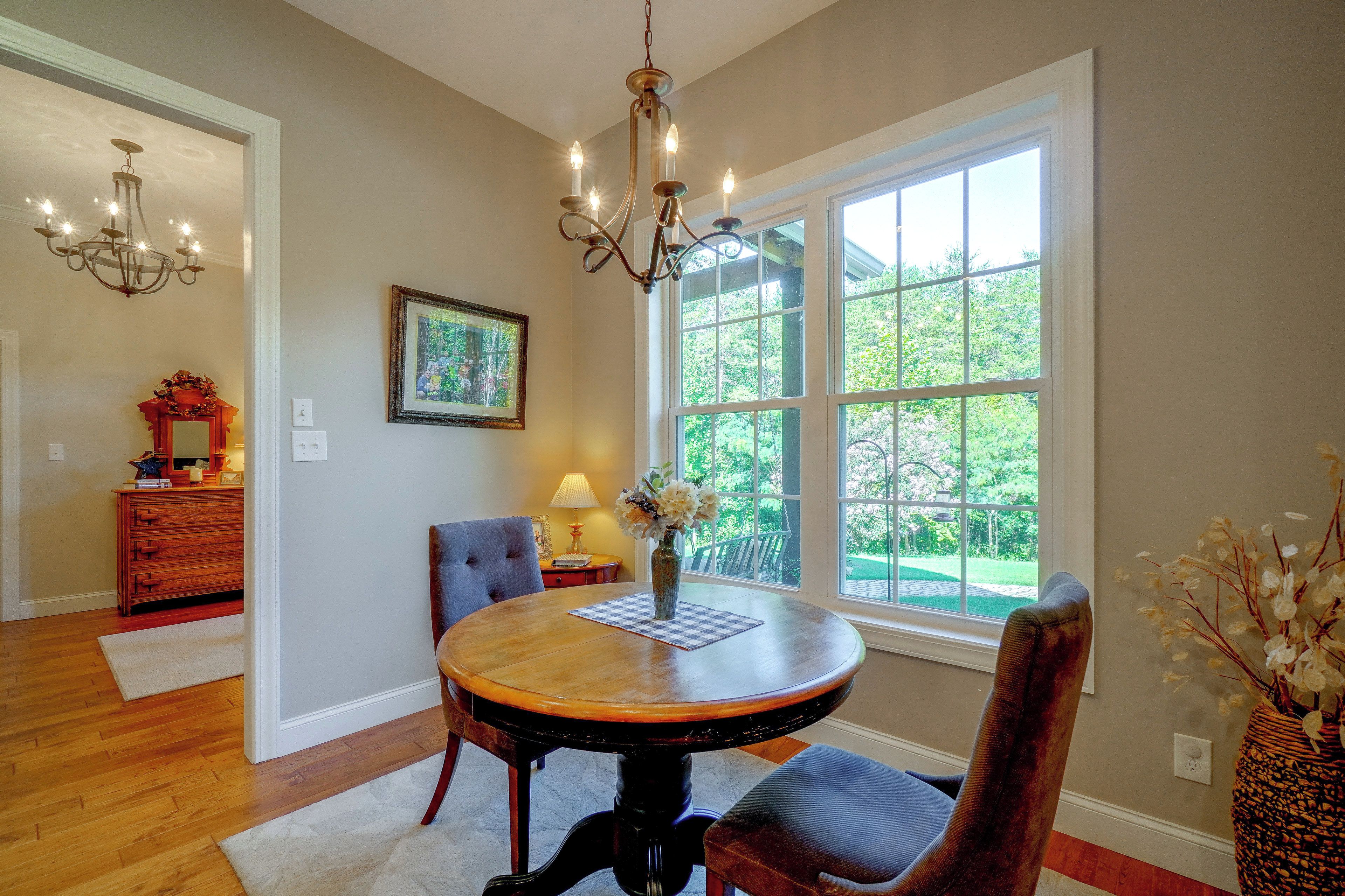 Une salle à manger avec une table ronde, des chaises, un lustre, une fenêtre donnant sur un paysage verdoyant et un tableau encadré au mur.