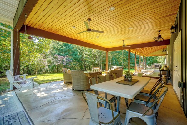Um alpendre coberto ao ar livre com mesa e cadeiras, um balanço e vista para um jardim.