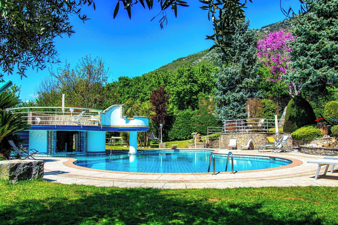 Ein Swimmingpool mit einer blauen Konstruktion, umgeben von viel Grün und einer Hügelkulisse im Hintergrund.