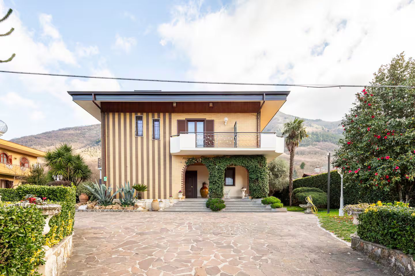 A two-story house with a stone-paved entrance, a balcony, and a garden with various plants and flowers.