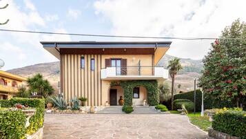 A two-story house with a stone-paved entrance, a balcony, and a garden with various plants and flowers.